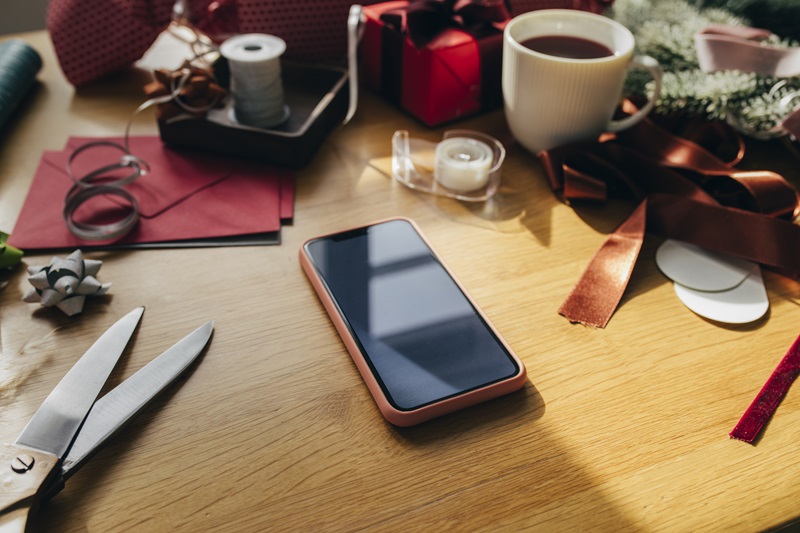 a smart phone a table surrounded by holiday Christmas presents, ribbons, ornaments, wreaths and other decorative items