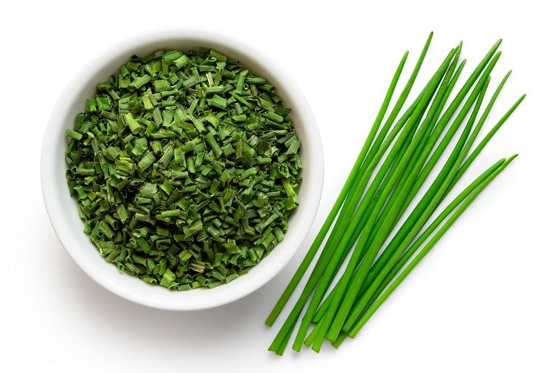 Dried chopped chives in white ceramic bowl next to a pile of fresh herbs